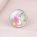 L'image représente une bague en métal avec un cabochon en verre. Le cabochon est rond et représente une licorne blanche avec une crinière et une queue roses. La licorne est entourée de fleurs et de feuilles.