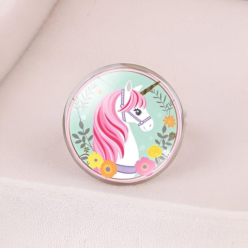 L'image représente une bague en métal avec un cabochon en verre. Le cabochon est rond et représente une licorne blanche avec une crinière et une queue roses. La licorne est entourée de fleurs et de feuilles.