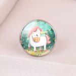L'image représente une bague en métal avec un cabochon en verre rond. Le cabochon est décoré d'une illustration représentant une licorne blanche et rose dans une forêt. La licorne est debout sur un pré vert et est entourée d'arbres et de fleurs.