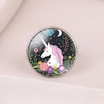 L'image représente une bague en métal argenté avec un cabochon en verre rond. Le cabochon est décoré d'une illustration colorée représentant une licorne blanche avec une crinière et une queue roses et violettes. La licorne est entourée de fleurs et de feuillage.