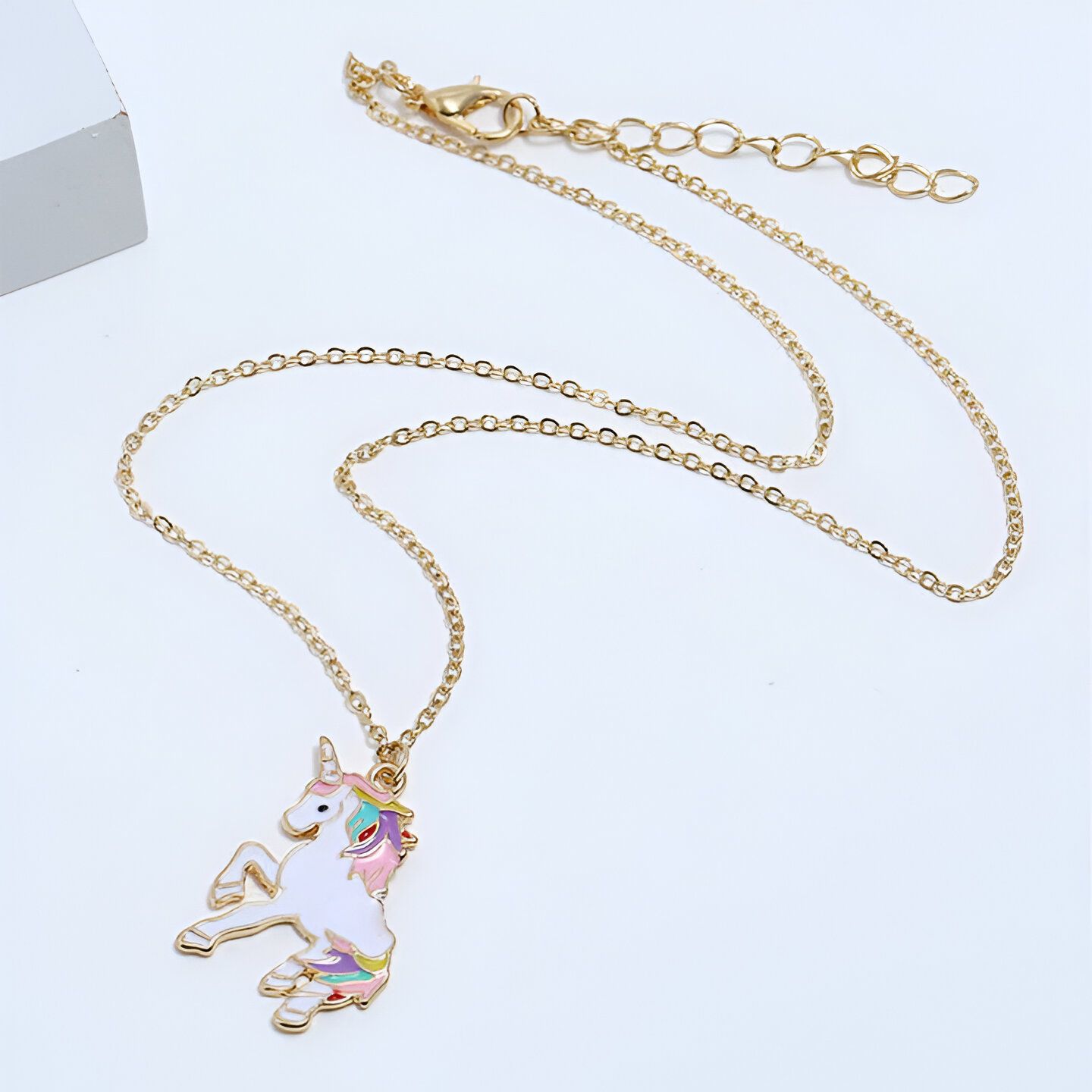 Un collier avec un pendentif licorne en émail coloré. La licorne est blanche avec une crinière et une queue arc-en-ciel. Elle est debout sur ses pattes arrière et a une expression joyeuse sur son visage. Le collier est en métal doré et la chaîne est réglable.