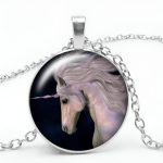 L'image représente un pendentif en forme de cheval blanc avec une corne sur le front. Le cheval est entouré d'un cercle en métal argenté. Le pendentif est accroché à une chaîne en métal argenté.