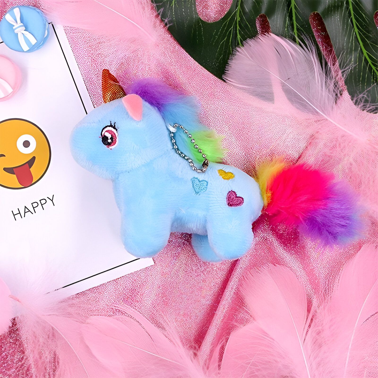 Une peluche licorne bleue avec une crinière et une queue arc-en-ciel. Elle est posée sur un fond rose avec des plumes blanches et un visage souriant en arrière-plan.